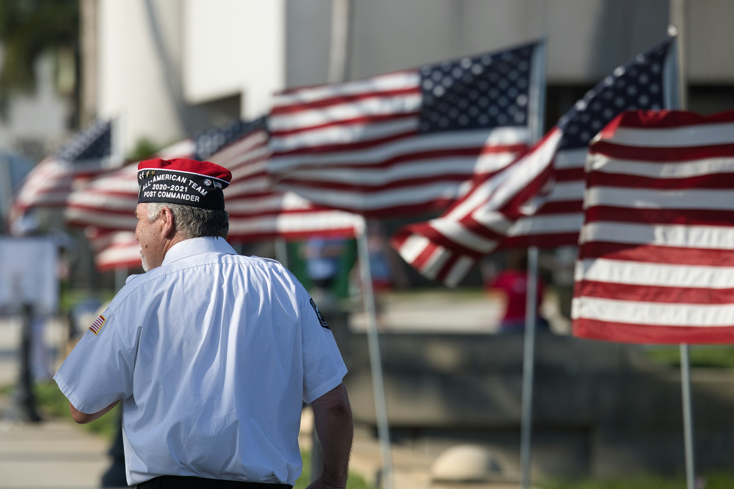 A veteran in a service cap standing before a row of American flags.